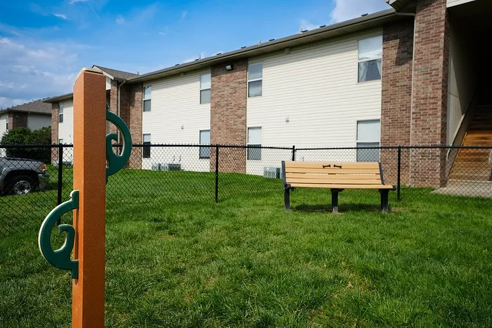 A pet-friendly area at Huntley Ridge East Apartments in Springfield, MO features a wooden bench and a green dog leash holder mounted on a brown post, all enclosed by a black chain-link fence on a well-maintained lawn.