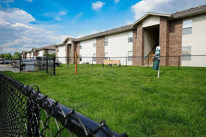 Wide-angle view of a fenced-in dog park area at Huntley Ridge East Apartments in Springfield, MO, showing a green pet waste station, wooden bench, and dog leash post, all set against a backdrop of brick apartment buildings and blue sky.