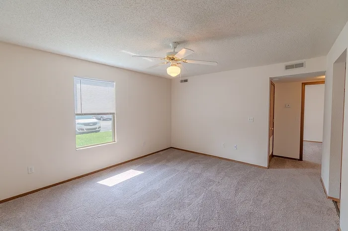 Carpeted living room at Huntley Ridge East Apartments in Springfield, MO with ceiling fan, natural light, and entry to hallway leading to bedrooms.