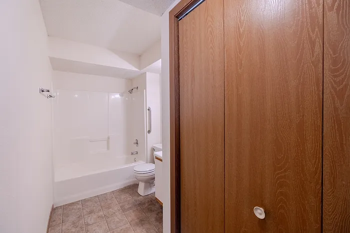 Bathroom layout at Huntley Ridge East Apartments in Springfield, MO showing a shower-tub combo, toilet, and white vanity with a wood-trimmed cabinet. A wooden door on the right provides access to in-bathroom storage space.