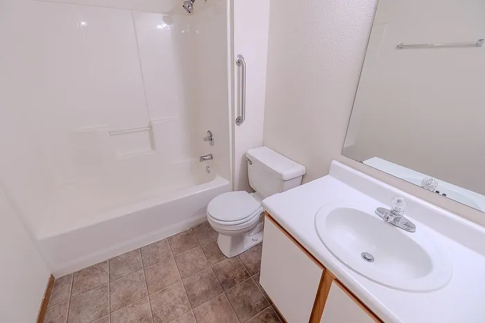 Modern bathroom at Huntley Ridge East Apartments in Springfield, MO featuring a white vanity with sink, toilet, and a clean shower-tub combo with built-in shelving and grab bar.