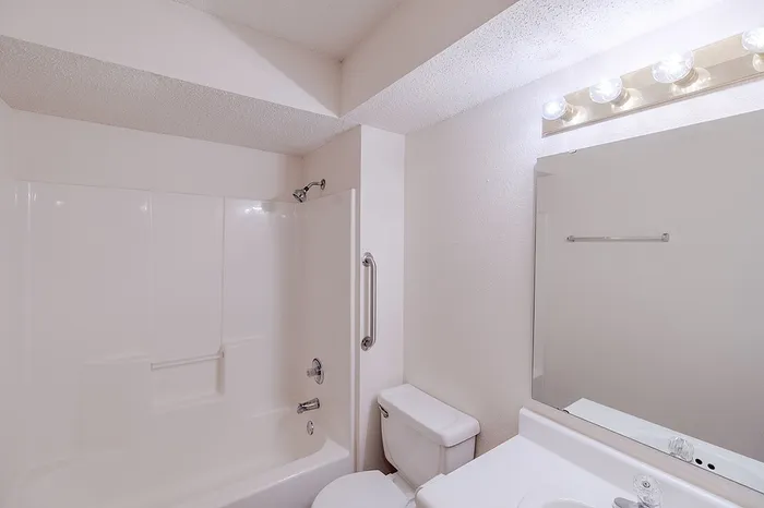 Bright bathroom view at Huntley Ridge East Apartments in Springfield, MO showcasing a fiberglass shower-tub combo, grab bar, and overhead vanity lighting above a large mirror.