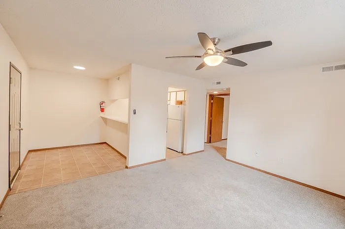Spacious living room at Huntley Ridge East Apartments in Springfield, MO, featuring neutral-tone carpeting, a modern ceiling fan, and open access to a tiled dining nook near the kitchen.