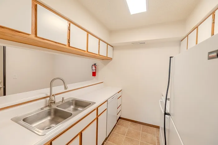 Bright and efficient galley kitchen at Huntley Ridge East featuring white countertops, a double stainless-steel sink, overhead cabinets with wood trim, and updated appliances.