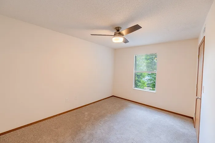 Bedroom inside a 1 bedroom unit at Huntley Ridge East, featuring plush carpeting, neutral walls, a modern ceiling fan, and window with leafy outdoor view.