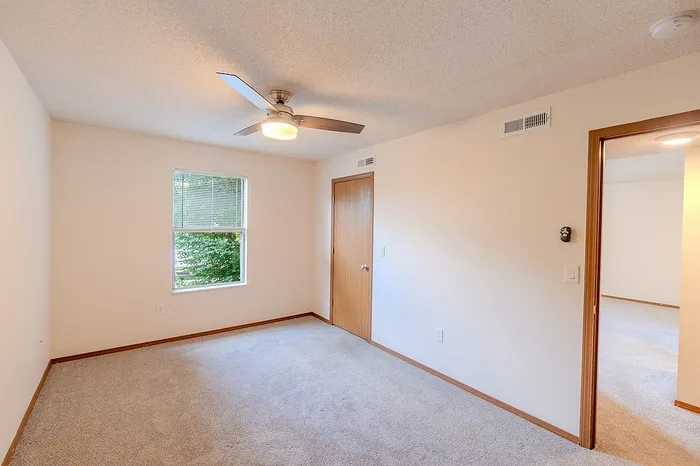 Alternate angle of the bedroom showing the entry to the hallway and closet, with ceiling fan and soft natural light from the single window.