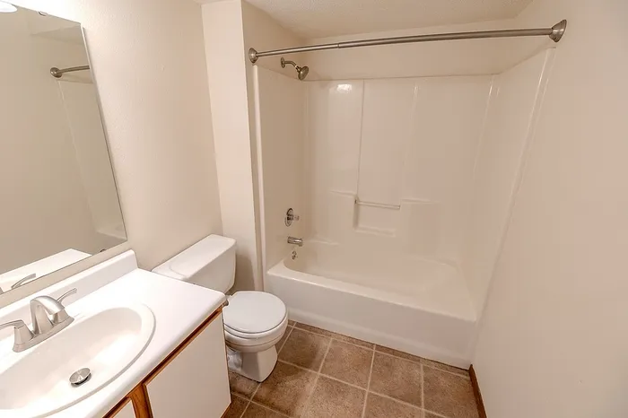 Bright bathroom at Huntley Ridge East featuring a white vanity with mirror, standard toilet, and full-sized shower/tub combo with built-in shelving and tile flooring.