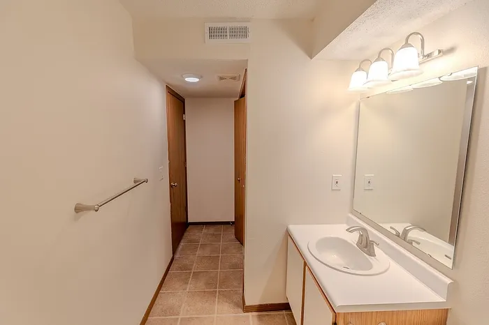 Wide-angle view of the upgraded bathroom showing the white sink vanity, wall-mounted towel bar, and tile flooring leading to the hallway and built-in storage area.
