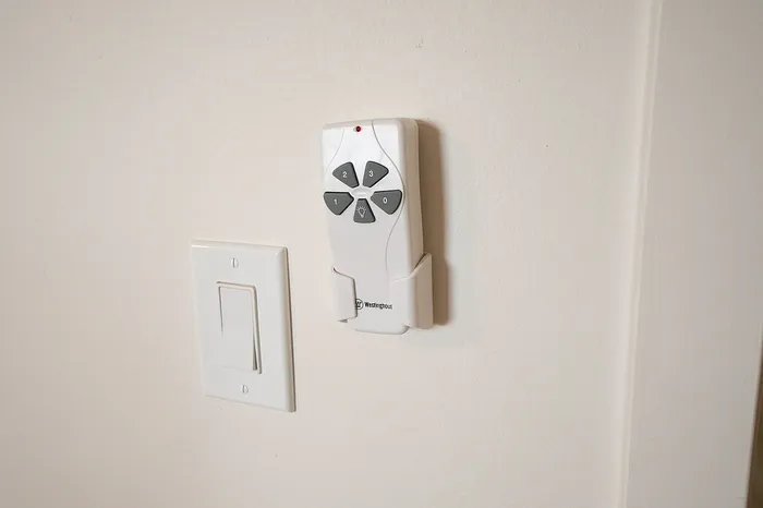 A close-up view of a Westinghouse ceiling fan remote control mounted on a beige wall, located next to a white light switch.