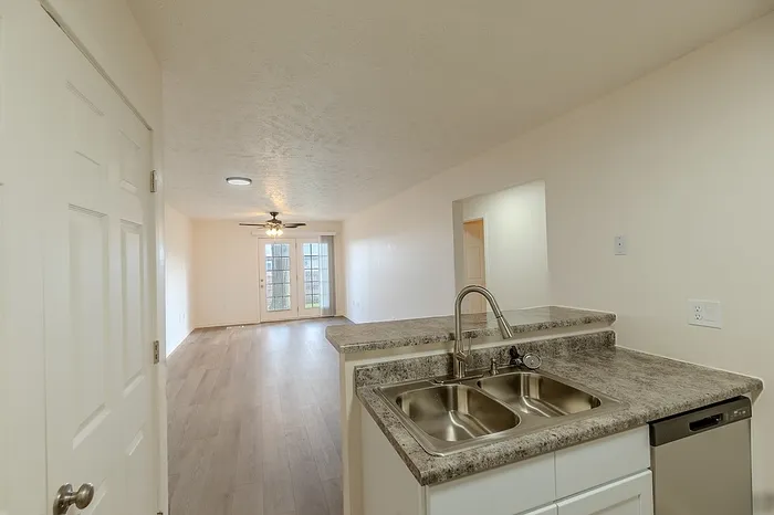 View from the kitchen showing a dual sink, raised bar counter, and spacious living room with wood-style flooring and natural light from the back doors.