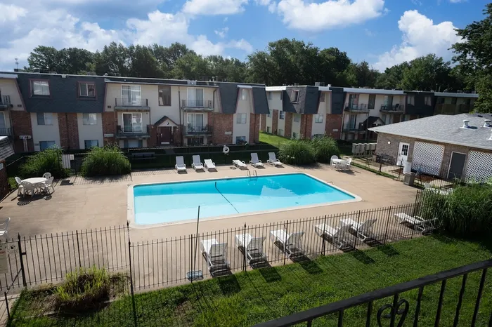 Fenced-in outdoor swimming pool at Rosewood Village Apartments in Springfield, MO, surrounded by lounge chairs and patio seating, with two-story apartment buildings and a clubhouse in the background under a partly cloudy sky.
