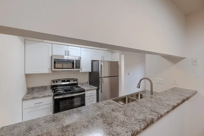 Modern kitchen at Huntley Ridge East in Springfield, MO, featuring stainless steel appliances, white cabinetry, a double sink, and a raised breakfast bar with granite-style countertops.
