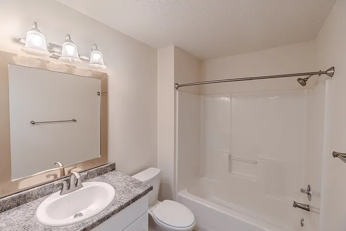 Modern bathroom at Huntley Ridge East apartments, featuring a large mirror with decorative lighting, single-sink vanity with granite-style countertop, and a full tub and shower combination.