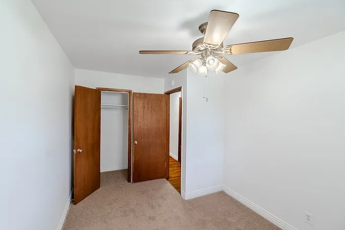 Bedroom at Village Club Apartments in Springfield, MO, featuring soft carpet, white walls, ceiling fan with lights, a window for natural light, and a reach-in closet for storage.