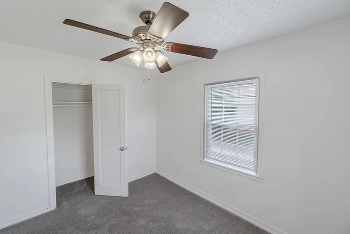 Bedroom at Village Club Apartments in Springfield, MO, with a ceiling fan, gray carpet, and window letting in natural light.