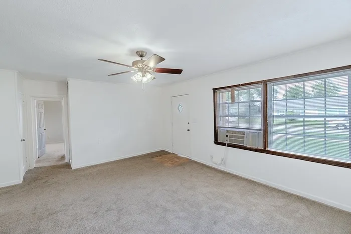 Living room at Village Club Apartments in Springfield, MO, featuring large windows, a ceiling fan, beige carpet, and front door entry.