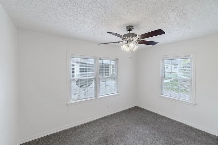 Bedroom at Village Club Apartments in Springfield, MO, with two large windows, gray carpet, and a ceiling fan providing overhead light.