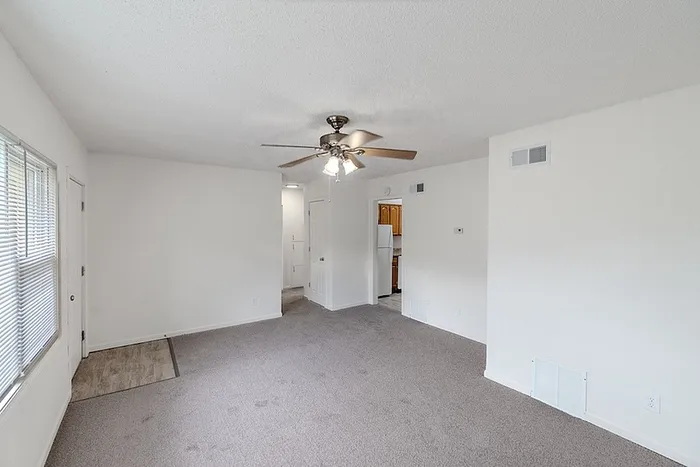 Open living area at Village Club Apartments in Springfield, MO, with neutral walls, ceiling fan, and view into the kitchen and hallway.