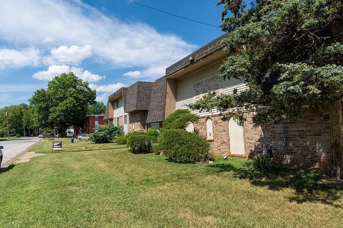 Charming suburban apartment complex with lush green lawn and mature trees, offering a serene living environment under a bright blue sky.
