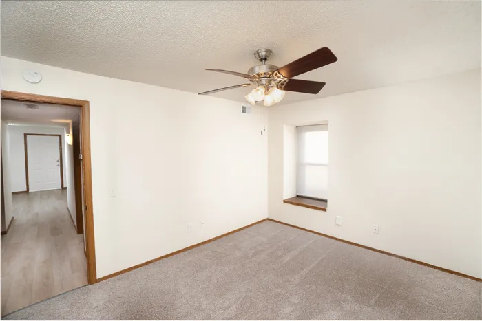 Bedroom at Catalpa East Apartments in Springfield, MO, with carpeted flooring, ceiling fan, window, and doorway leading to the interior hallway.