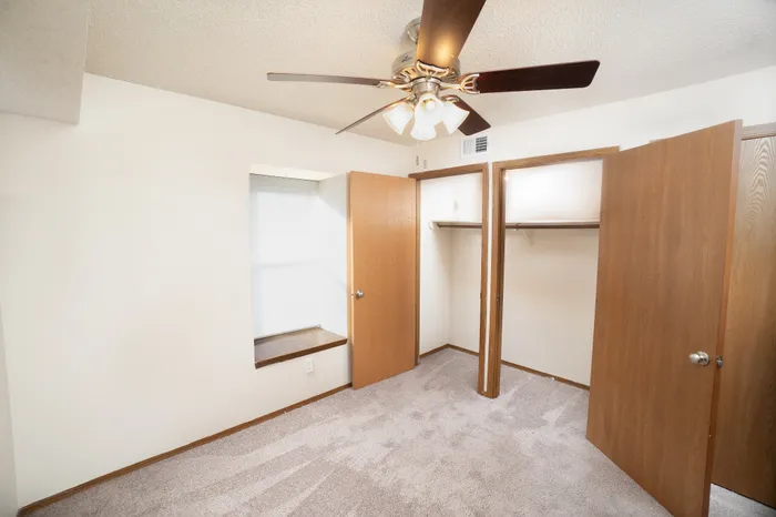 Bedroom at Catalpa East Apartments in Springfield, MO, featuring carpeted flooring, neutral walls, a ceiling fan with light, a window with sill, and an open closet with shelving.