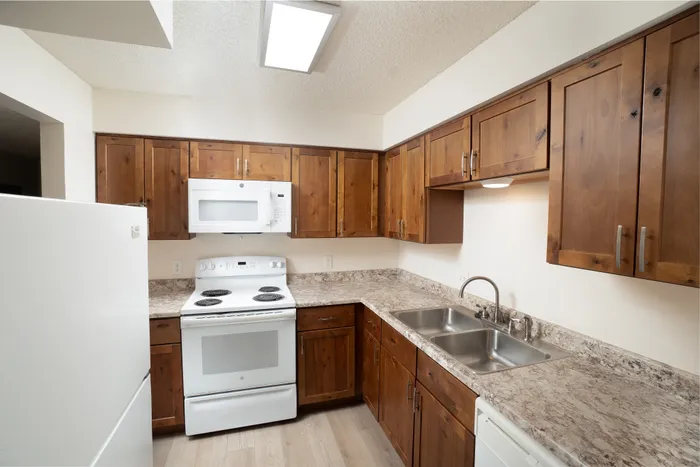 Kitchen at Catalpa East Apartments in Springfield, MO, with wood cabinetry, laminate countertops, white appliances including stove and microwave, and a double-basin sink.
