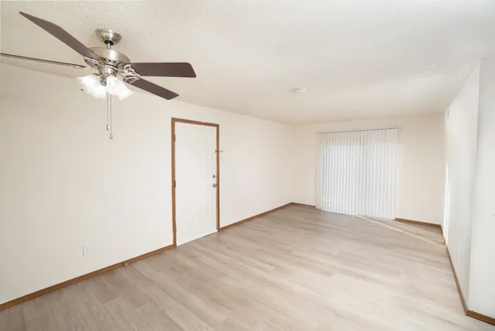 Bright living room at Catalpa East Apartments in Springfield, MO, featuring light wood-style flooring, neutral walls, a ceiling fan with light fixture, and sliding glass doors with vertical blinds.ht, spacious living area with sleek wooden flooring and a modern ceiling fan, perfect for comfortable living.