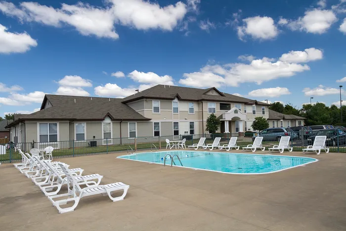 Outdoor swimming pool at Chesterfield Village Apartments with a concrete deck, white lounge chairs, and a gated fence, with two-story apartment buildings in the background under a partly cloudy sky.