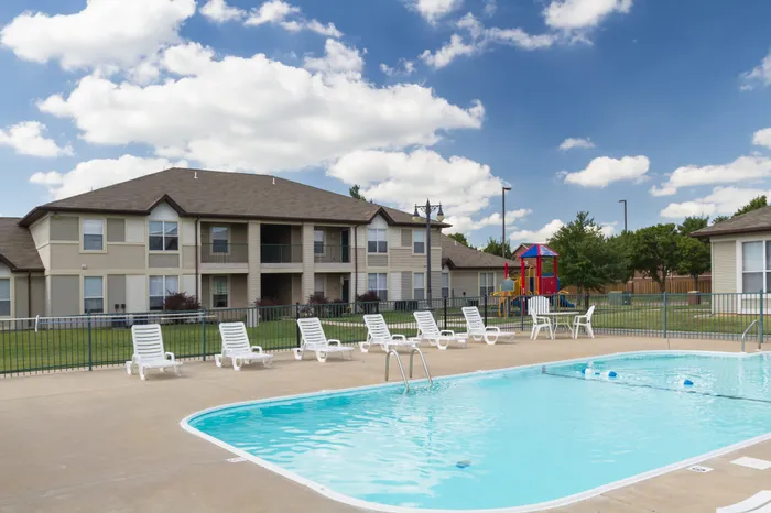 Chesterfield Village community pool with white lounge chairs, grassy courtyard, apartment buildings in the background, and a colorful playground nearby.