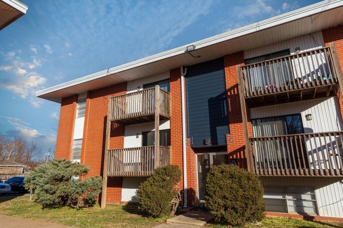 Front exterior of a Seminole North Apartments building with brick fa&ccedil;ade, vertical blue accent panel, stacked wooden balconies, ground-level entry, and trimmed shrubs along the walkway.