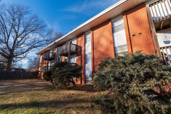 Angled side view of a two-story brick apartment building at Seminole North Apartments, featuring wood-railed balconies on the upper level, large vertical windows, trimmed evergreen shrubs, mature trees, and open lawn under a bright blue sky.