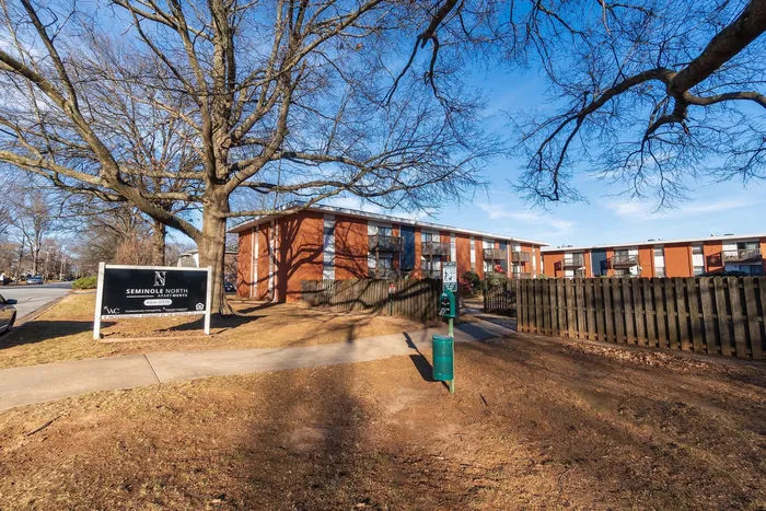 Exterior view of Seminole North Apartments in Springfield, Missouri, showing the community entrance sign along a sidewalk, a fenced walkway, mature shade trees, and two-story brick apartment buildings with private balconies set back from the street.