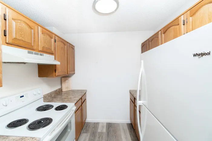 Full kitchen view with wood cabinetry, white refrigerator, electric range, laminate countertops, and durable wood-style flooring at Seminole North Apartments.