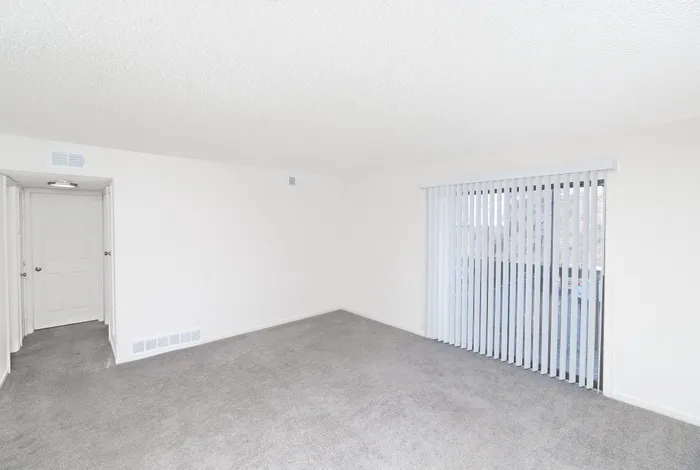 Bright living room with neutral carpet flooring, white walls, and a sliding glass door with vertical blinds leading to a private patio or balcony at Seminole North Apartments.