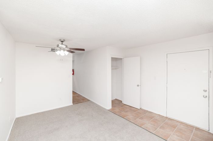 Bright apartment entry and living area with carpeted flooring, ceiling fan with light, tiled entryway, and an open closet near the front door.