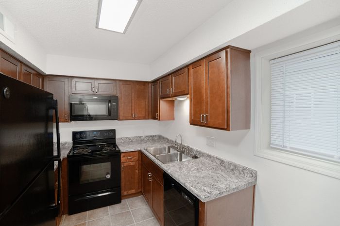 Compact kitchen with wood cabinetry, gray laminate countertops, double-basin sink, black appliances, tiled flooring, and a window with blinds.