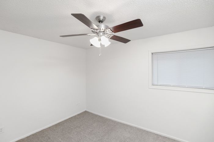 Carpeted bedroom with white walls, ceiling fan with light, and a window with blinds providing natural light.