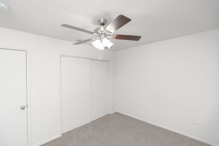 Bedroom view showing carpeted flooring, ceiling fan with light, sliding closet doors, and an entry door.