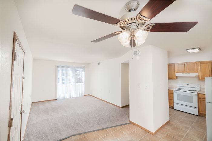 Bright and inviting living space with a ceiling fan, spacious carpeted area, and modern kitchen featuring wooden cabinets and white appliances.