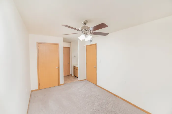 Bright, newly carpeted room with a modern ceiling fan, featuring wooden doors leading to a stylish bathroom area. Ideal for a cozy living space.