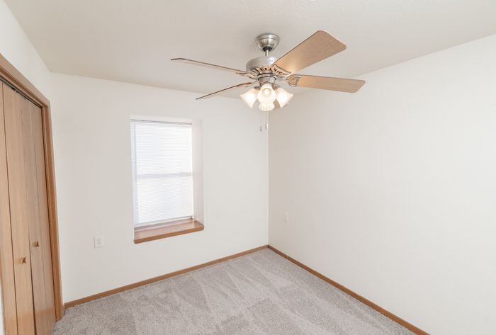 Bright and cozy room featuring soft carpet flooring, a wooden ceiling fan, and a large window with blinds.