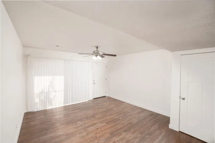 Bright living room with wood-style flooring, ceiling fan, white walls, and sliding glass door with vertical blinds.