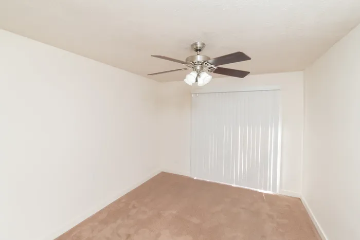 Bedroom with carpet flooring, ceiling fan, white walls, and sliding glass door with blinds.
