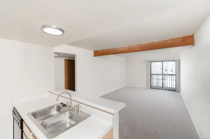 Living room with open kitchen pass-through, neutral carpet, ceiling beam detail, sliding patio door with vertical blinds