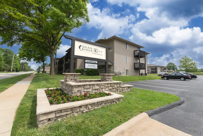 Charming apartment complex surrounded by lush greenery and well-maintained landscaping, with ample parking space under a vibrant blue sky.