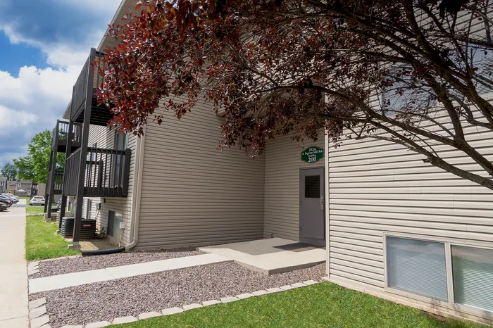 Inviting apartment entrance with modern siding, lush green surroundings, and quaint balconies under a vibrant blue sky.