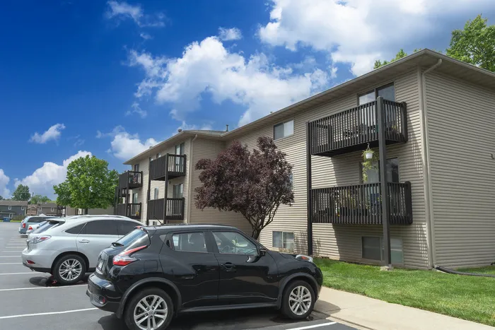 Charming apartment complex with inviting balconies, surrounded by lush greenery and a vibrant blue sky, complemented by a convenient parking area.