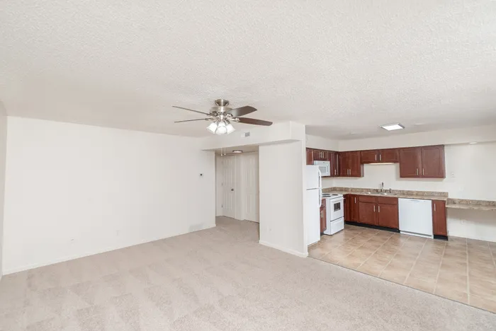 Open living area with carpet, ceiling fan, and adjacent kitchen at Quail Ridge East Apartments.