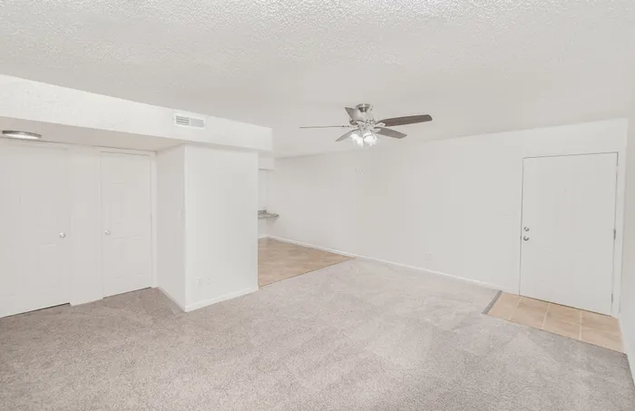 Spacious living room with carpet, entry door, ceiling fan, and open kitchen view at Quail Ridge East Apartments.