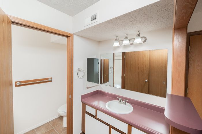 Bathroom vanity with laminate countertop, large mirror, lighting fixture, and adjacent toilet area.